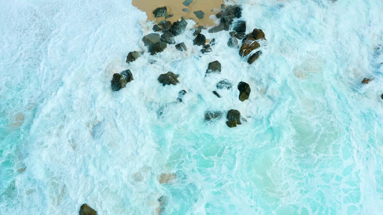 Aerial frame from Marquis Los Cabos with the main dark rock cluster more centered as white foam drains into turquoise water, with no visible people.