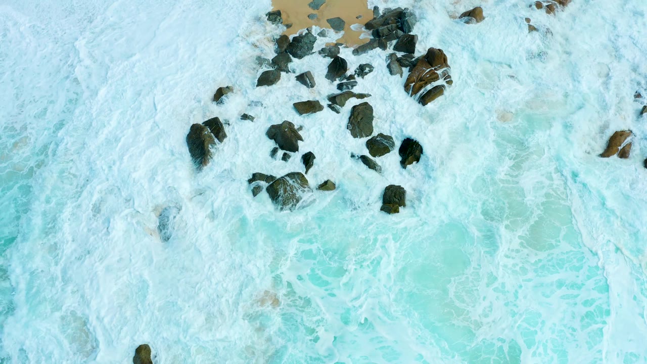 Top-down Marquis Los Cabos shoreline frame showing dense white foam, pale blue-green water, tan sand along the upper edge, and dark rocks with no visible people.