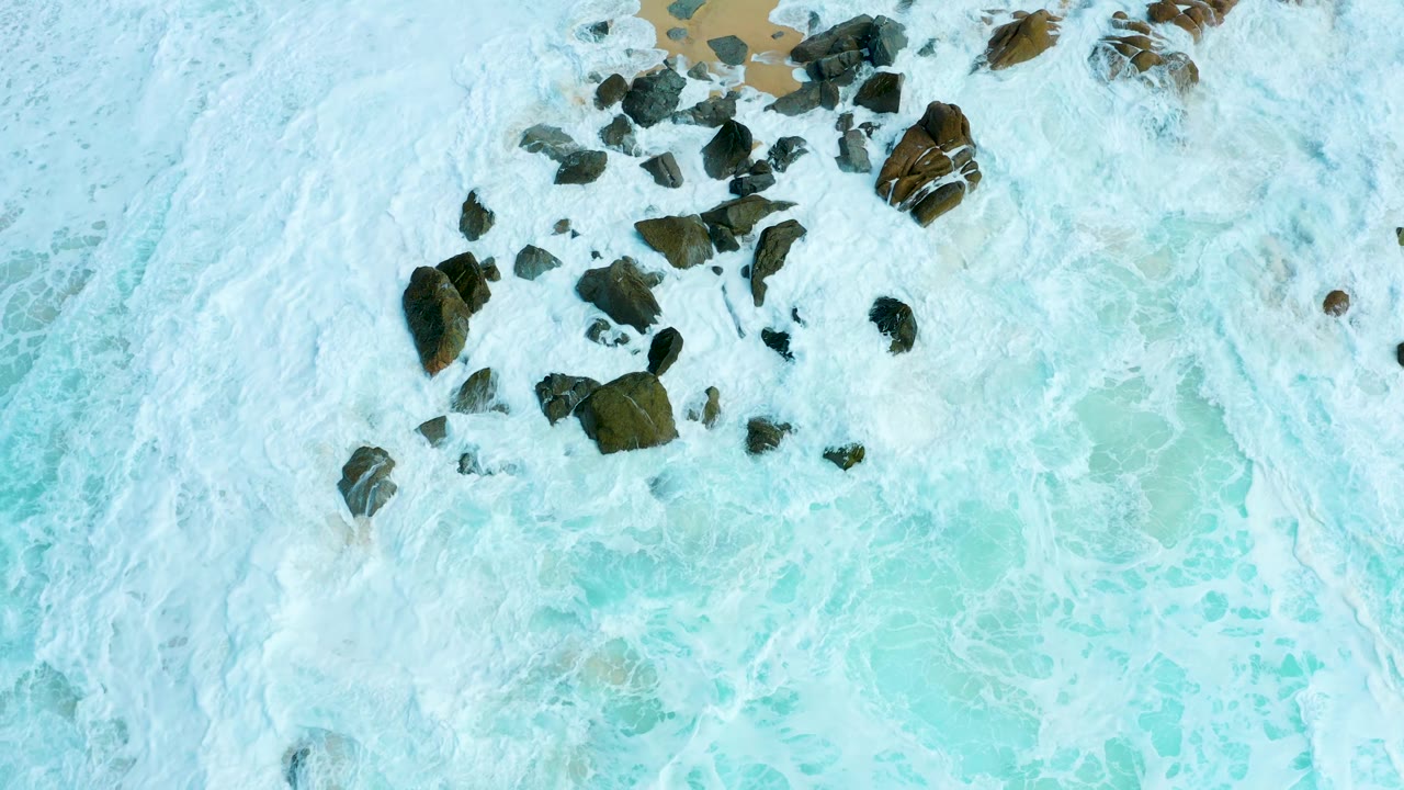Aerial view at Marquis Los Cabos where more of the dark rocks are visible as the white foam recedes across turquoise water, with no visible people.