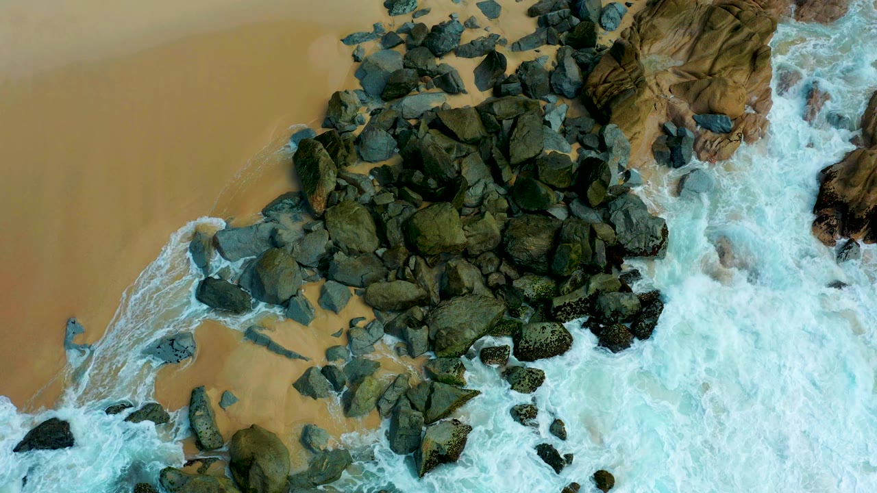 Overhead view of rocks, sand, and surf on a shoreline.