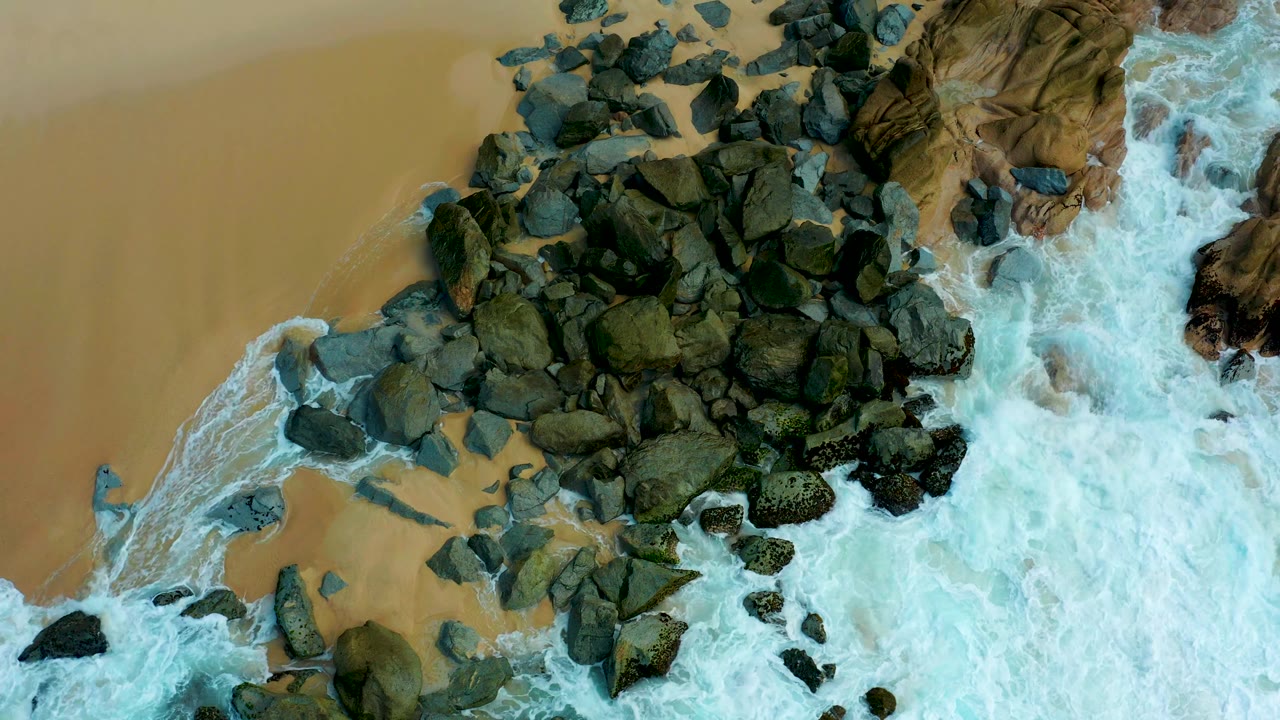 Aerial frame at Marquis Los Cabos with white foam wrapping around the lower rocks while tan sand remains visible at the upper left, with no visible people.