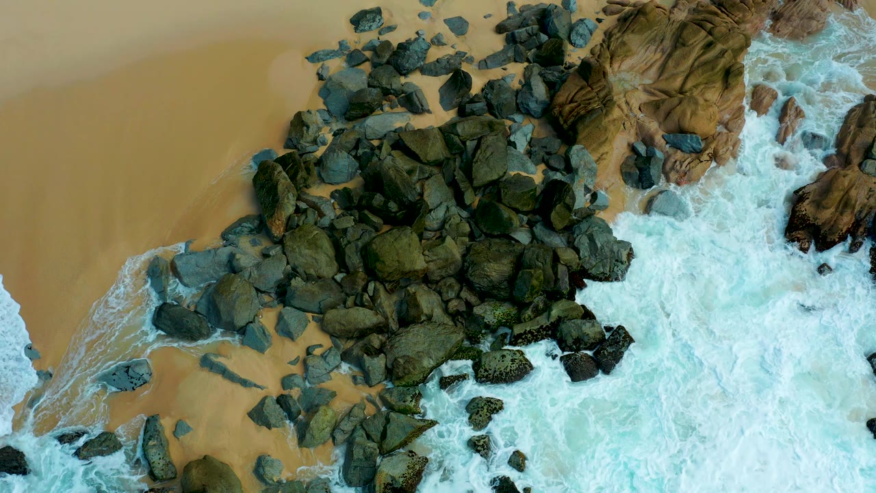 Aerial view of Marquis Los Cabos rocks and beach sand as bright foam spreads farther along the lower edge of the frame, with no visible people.