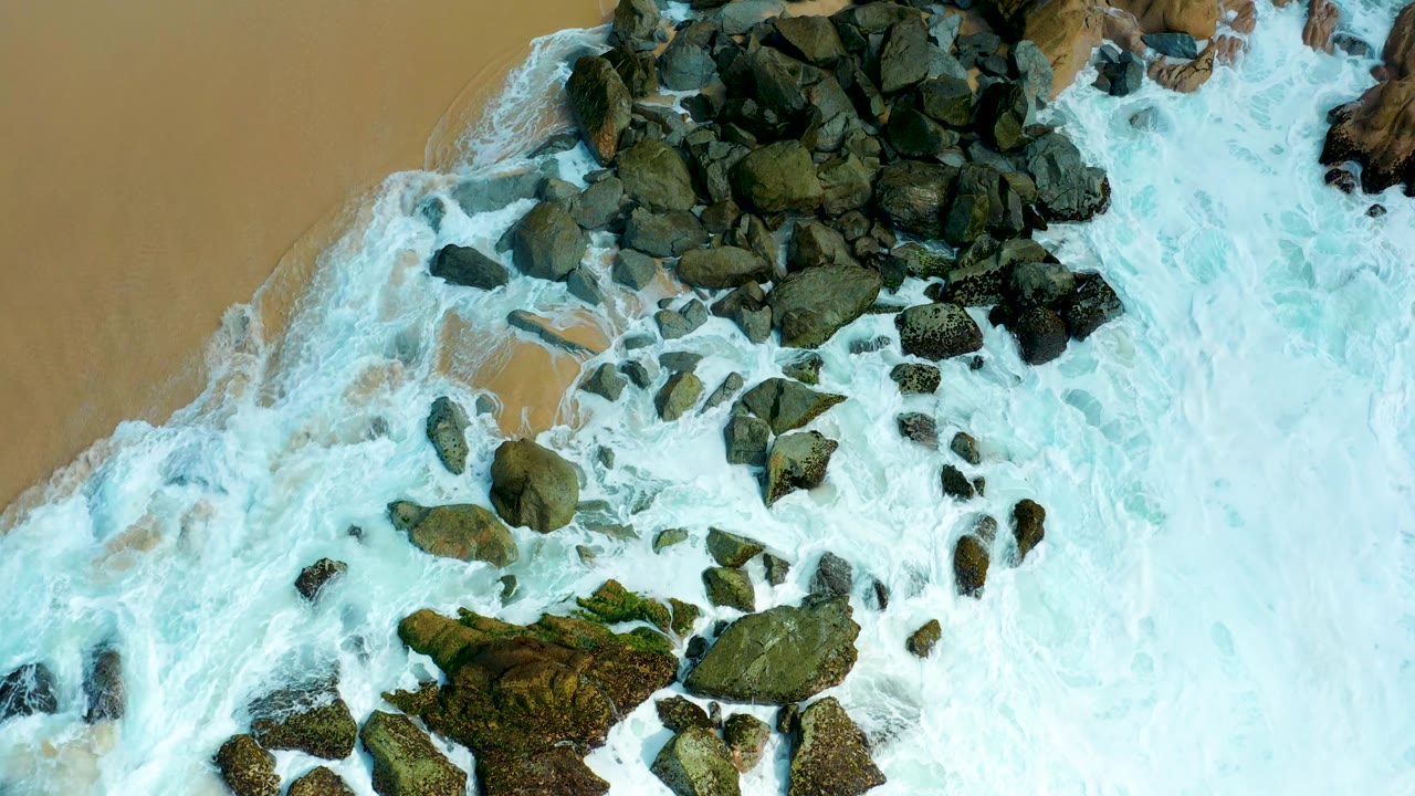 At 0:01, foamy blue-green water covers more of the central rocks while tan sand remains visible along the upper-left edge at Marquis Los Cabos, with no visible people.