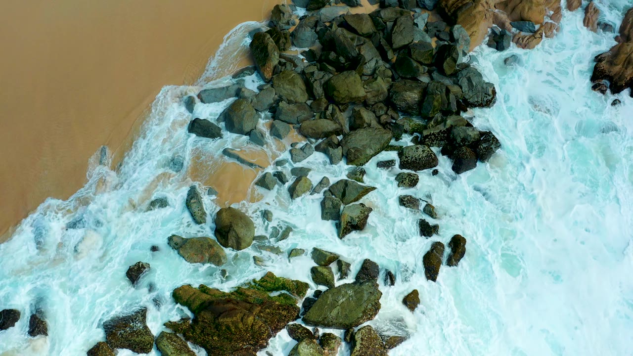 At 0:02, a diagonal sheet of white surf pushes around scattered dark rocks and tan beach at Marquis Los Cabos, with no visible people.