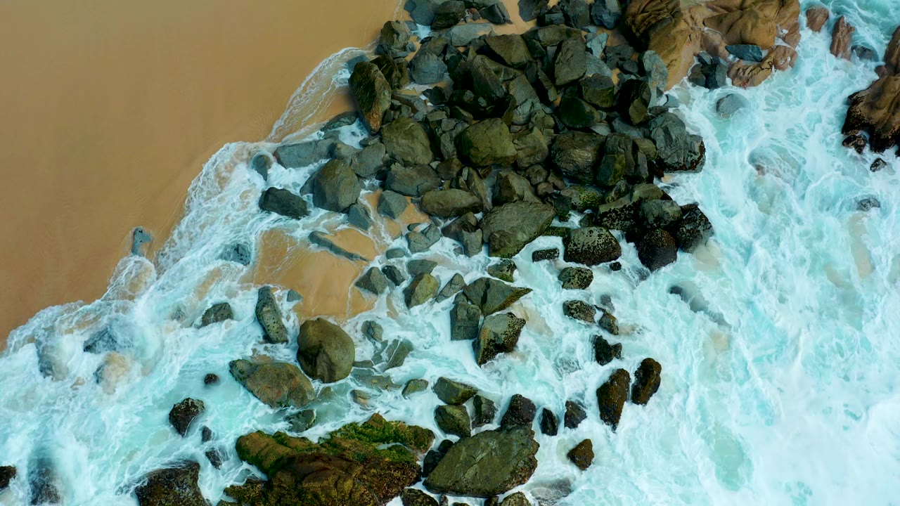 At 0:03, more of the dark rock cluster is visible through receding white water along the tan sand at Marquis Los Cabos, with no visible people.