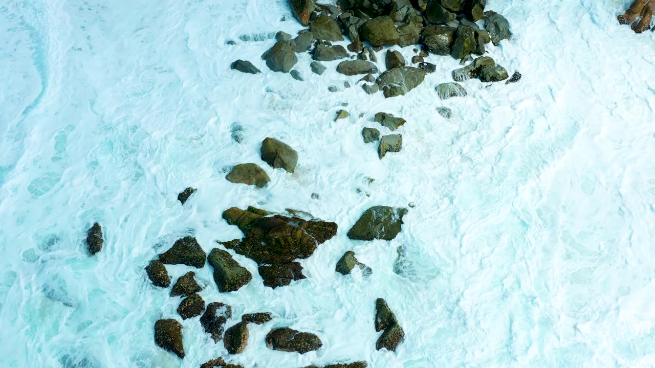 White and pale blue surf wraps around clustered rocks at Marquis Los Cabos, with the sandy shore still mostly hidden and no visible people.