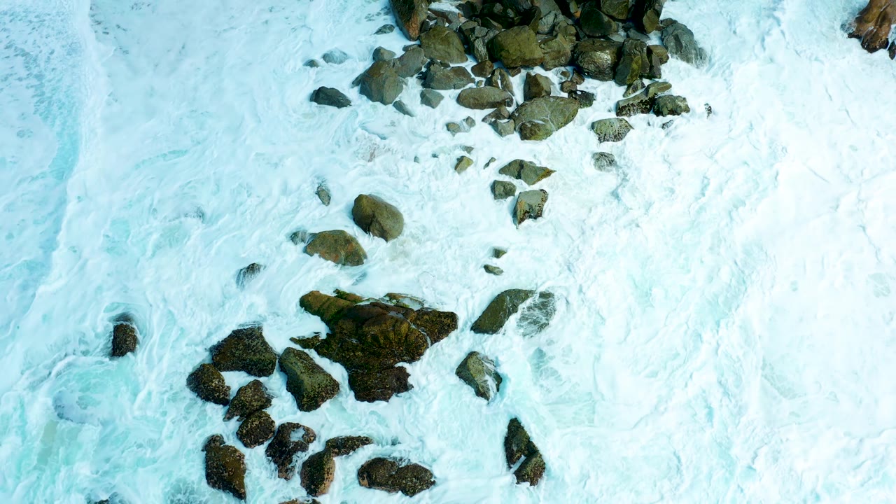 Aerial shoreline detail at Marquis Los Cabos shows streaked white foam, dark rocks, and a small tan sand edge entering the frame, with no visible people.