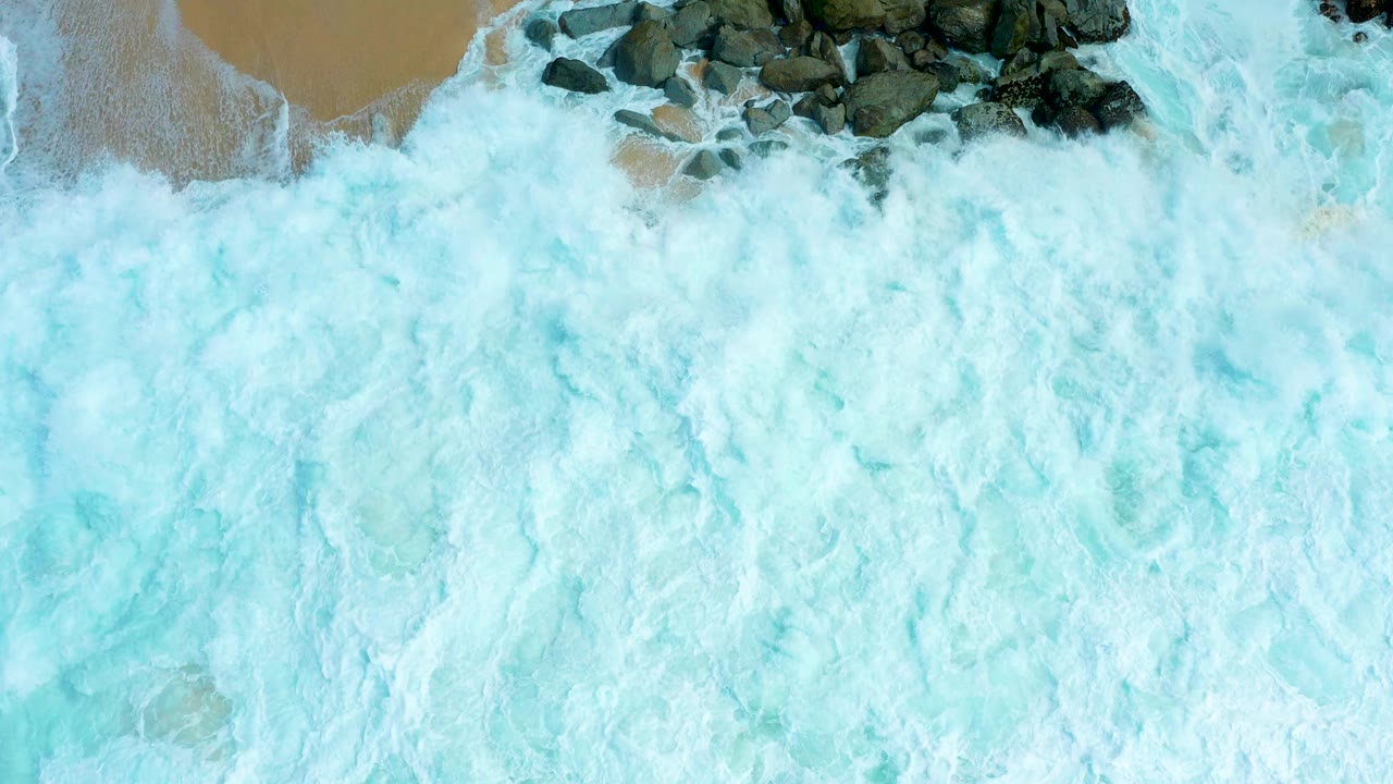 Overhead frame at Marquis Los Cabos shows white turquoise surf spreading across dark shoreline rocks with tan sand visible along the top edge and no visible people.