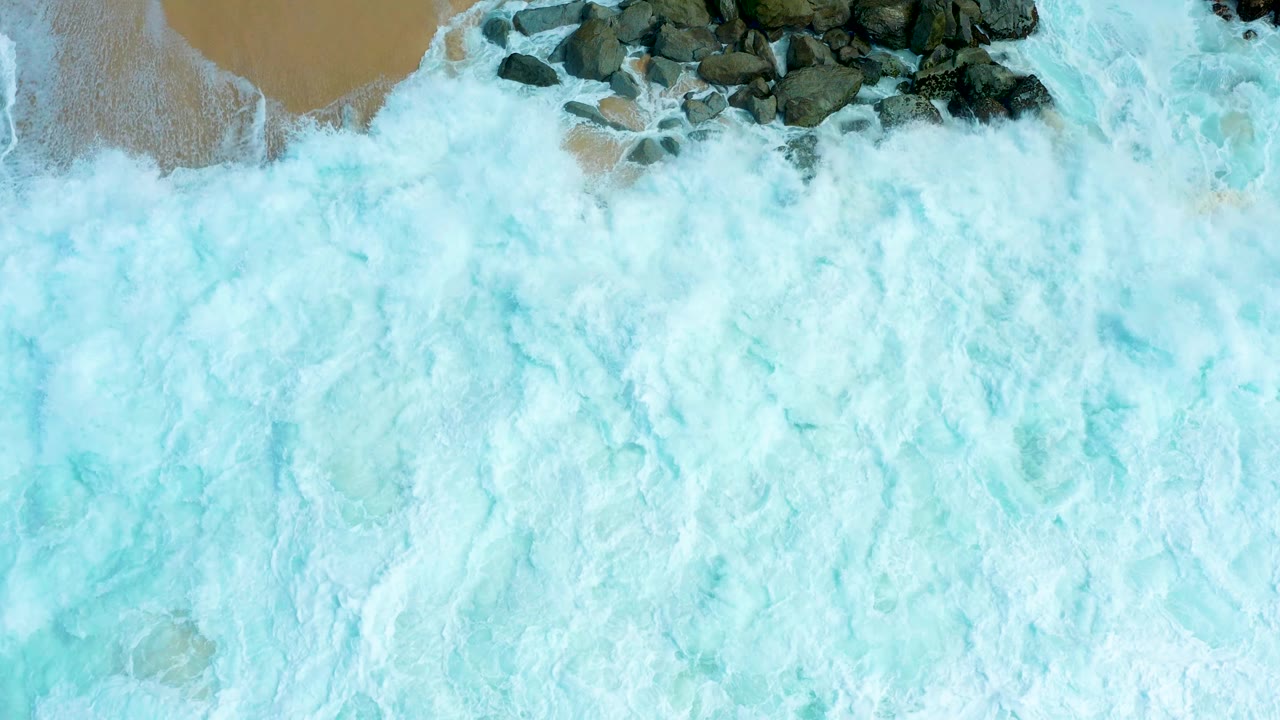 Overhead Marquis Los Cabos surf view with pale blue-white foam covering most of the water and a cluster of dark rocks near the upper edge, with no visible people.