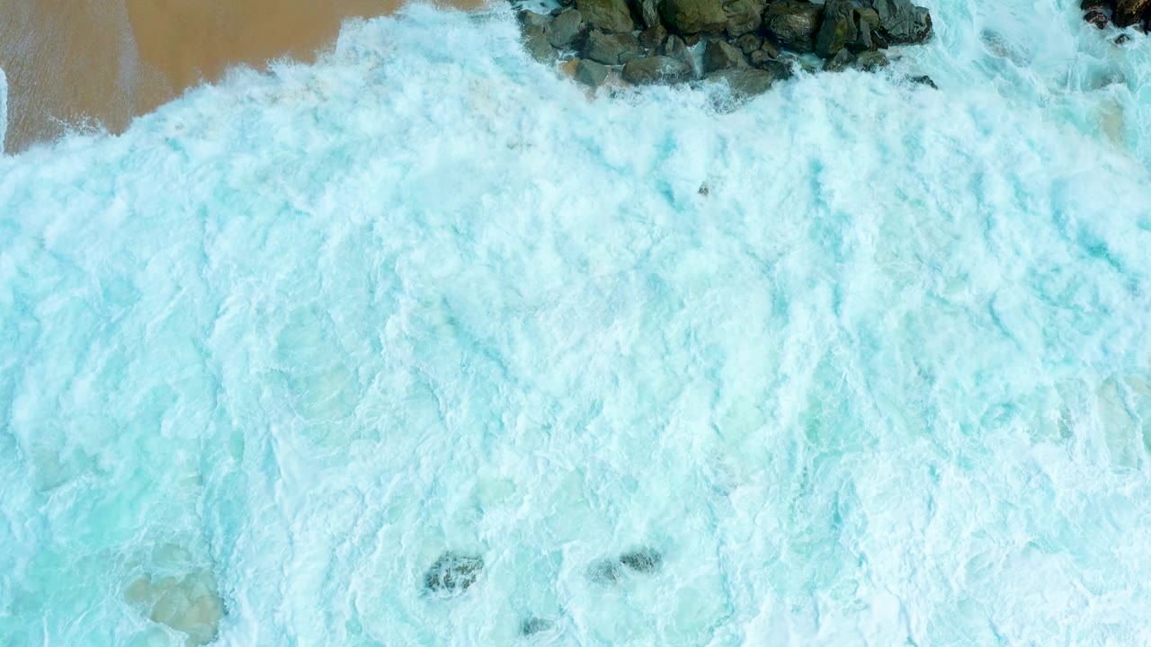 Aerial view at Marquis Los Cabos with turbulent white foam blanketing the shoreline and only small dark rocks breaking through the surf, with no visible people.