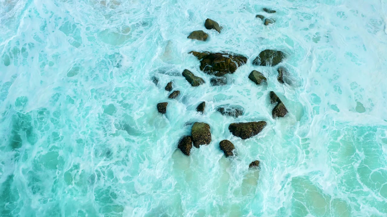 Aerial frame from Marquis Los Cabos with white foam spreading across aqua shallows and dark rocks grouped near the upper center, with no visible people.