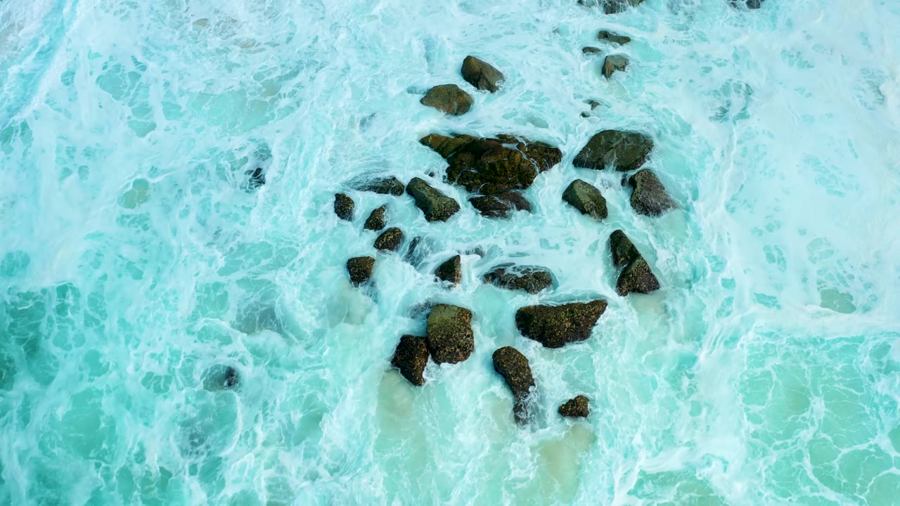 Top-down view at Marquis Los Cabos where a denser patch of white surf washes over dark rocks set in turquoise shallow water, with no visible people.