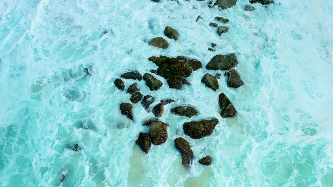 Aerial frame from Marquis Los Cabos showing a larger exposed rock group surrounded by pale turquoise water and lace-like foam, with no visible people.