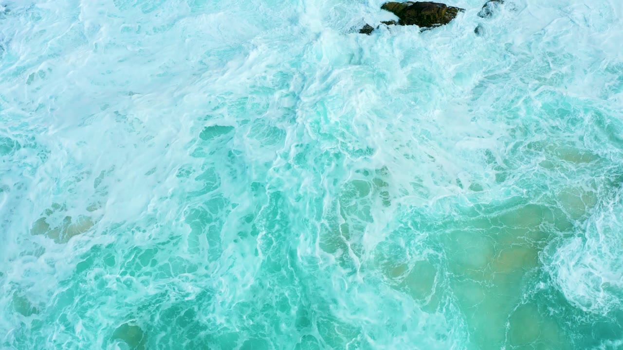 Top-down Marquis Los Cabos surf frame with white foam spreading across turquoise water and a dark rock set near the upper center, with no visible people.