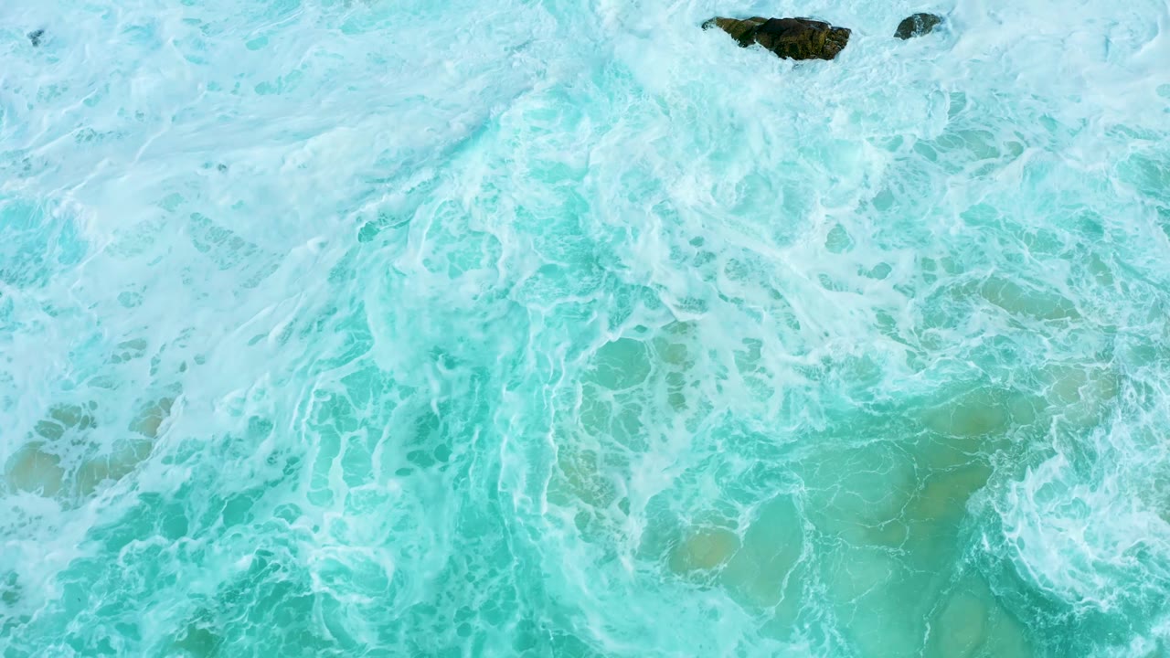 Overhead ocean view from Marquis Los Cabos with dense whitewater across turquoise sea and a dark rock near the upper right, with no visible people.