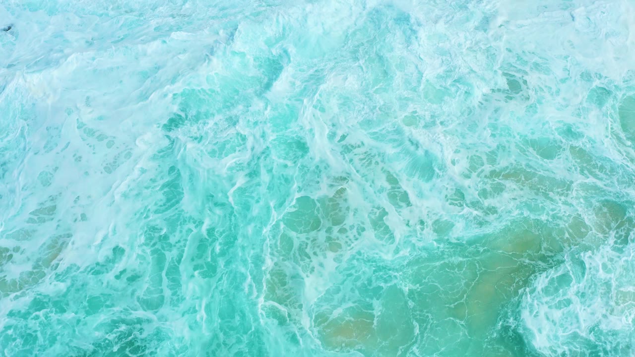 Top-down surf at Marquis Los Cabos where white foam thins over turquoise water and pale submerged patches show through, with no visible people.
