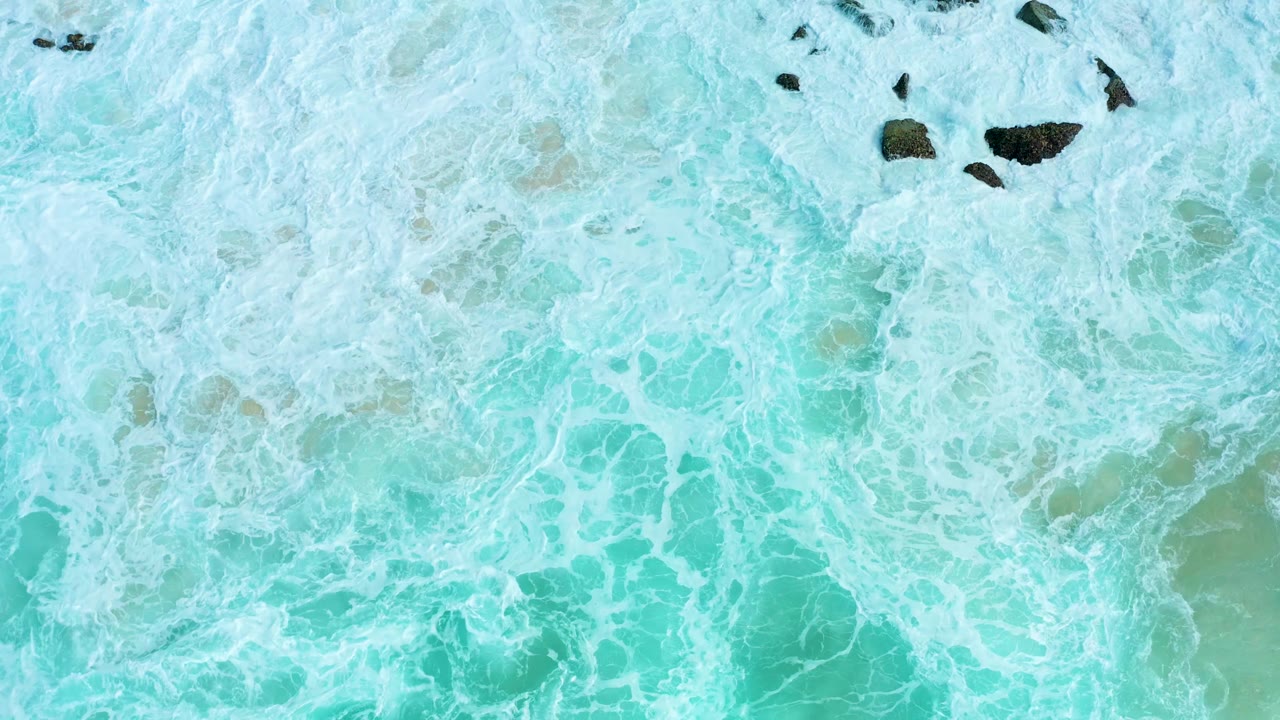 Blue-green ocean water at Marquis Los Cabos is covered by white foam patterns with a few dark rocks near the upper edge and no visible people.