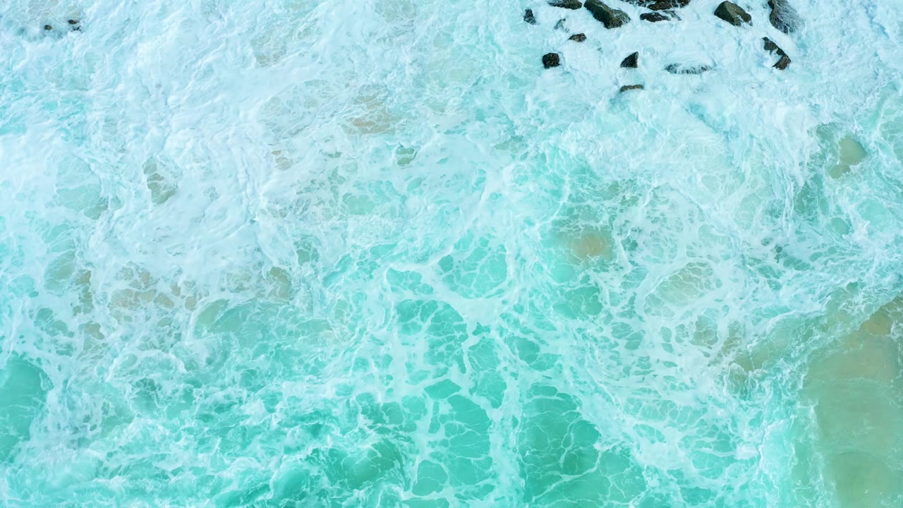 At Marquis Los Cabos, an overhead frame shows dense white surf over turquoise water with dark rocks clustered near the top and no visible people.