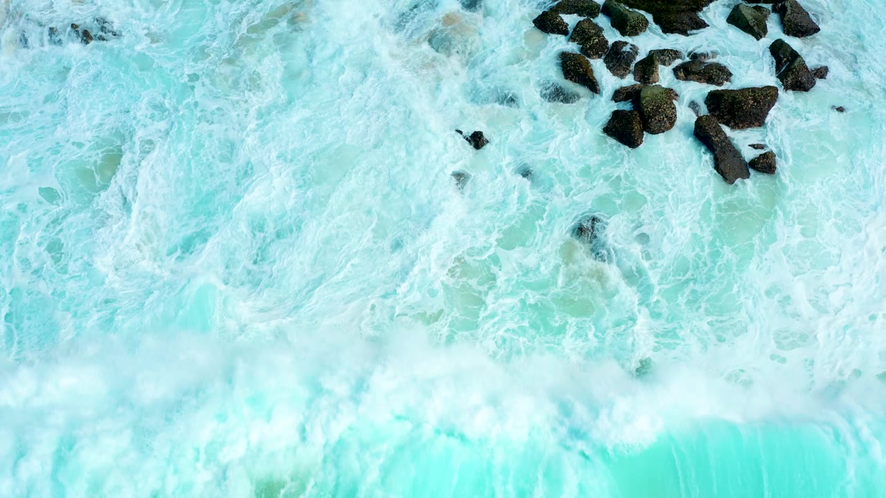 Turquoise surf and white foam around dark shoreline rocks.