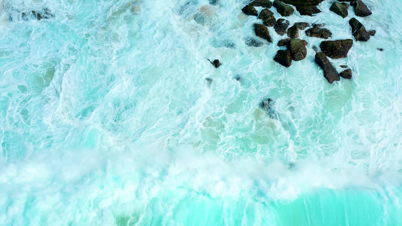Whitewater spreads across bright turquoise water near a clustered group of dark rocks at Marquis Los Cabos, Mexico, with no visible people.
