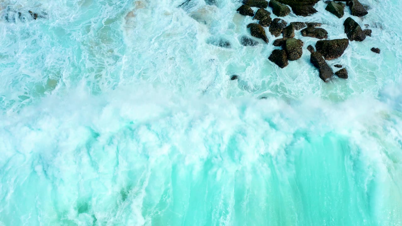 A dense band of white foam crosses pale turquoise surf while dark rocks remain partly exposed at Marquis Los Cabos, Mexico, with no visible people.