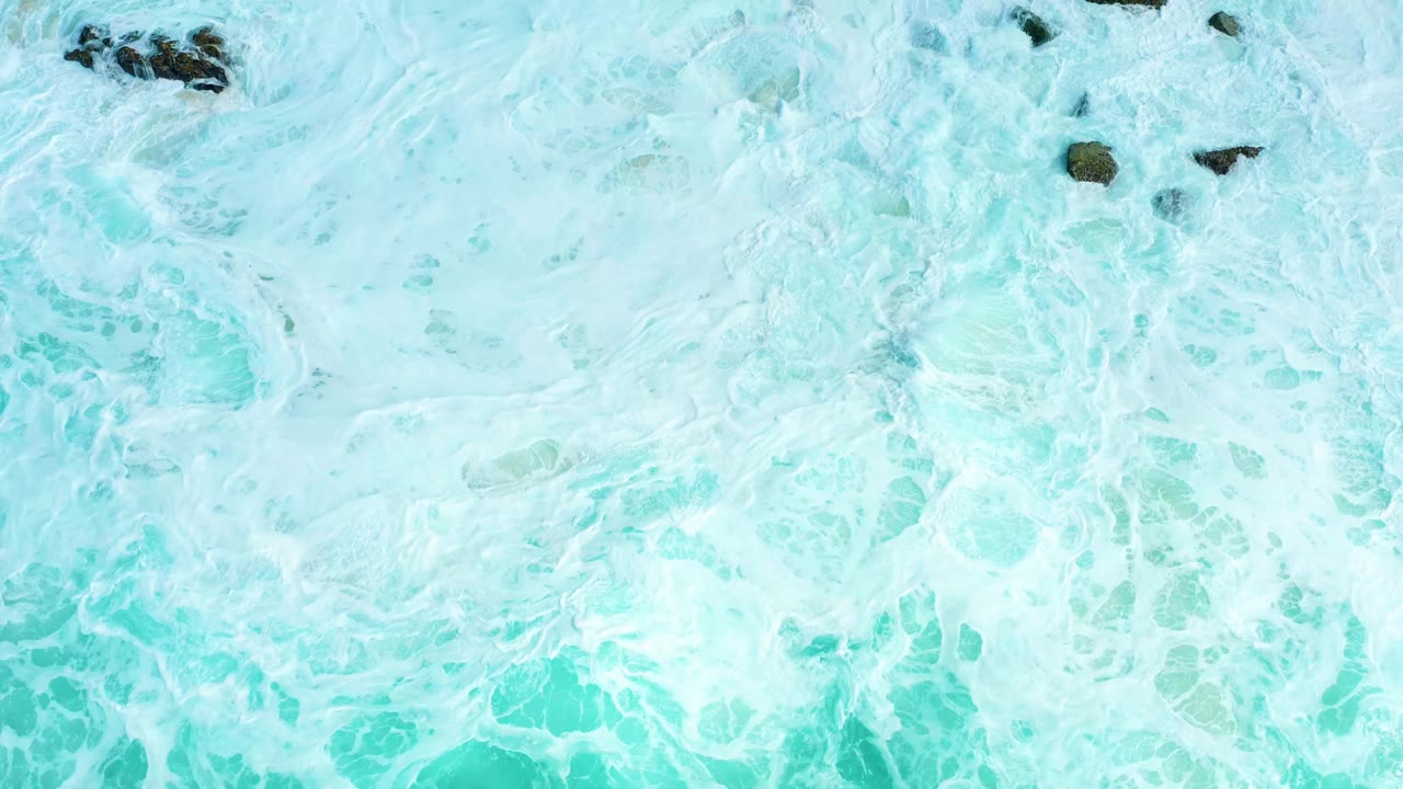 Aerial frame from Marquis Los Cabos with white foam veining across turquoise water and a small cluster of dark rocks near the upper edge, with no visible people.