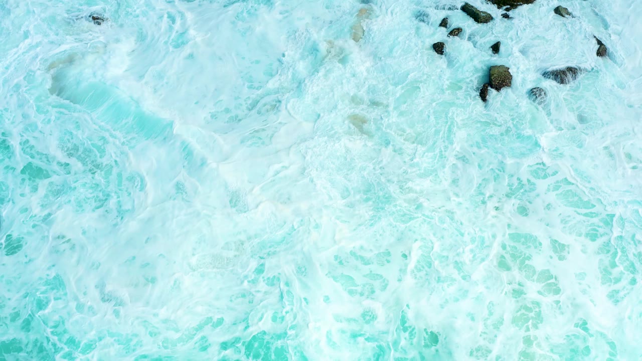 Overhead view at Marquis Los Cabos where a larger cluster of dark rocks sits in bright foamy turquoise surf, with no visible people.