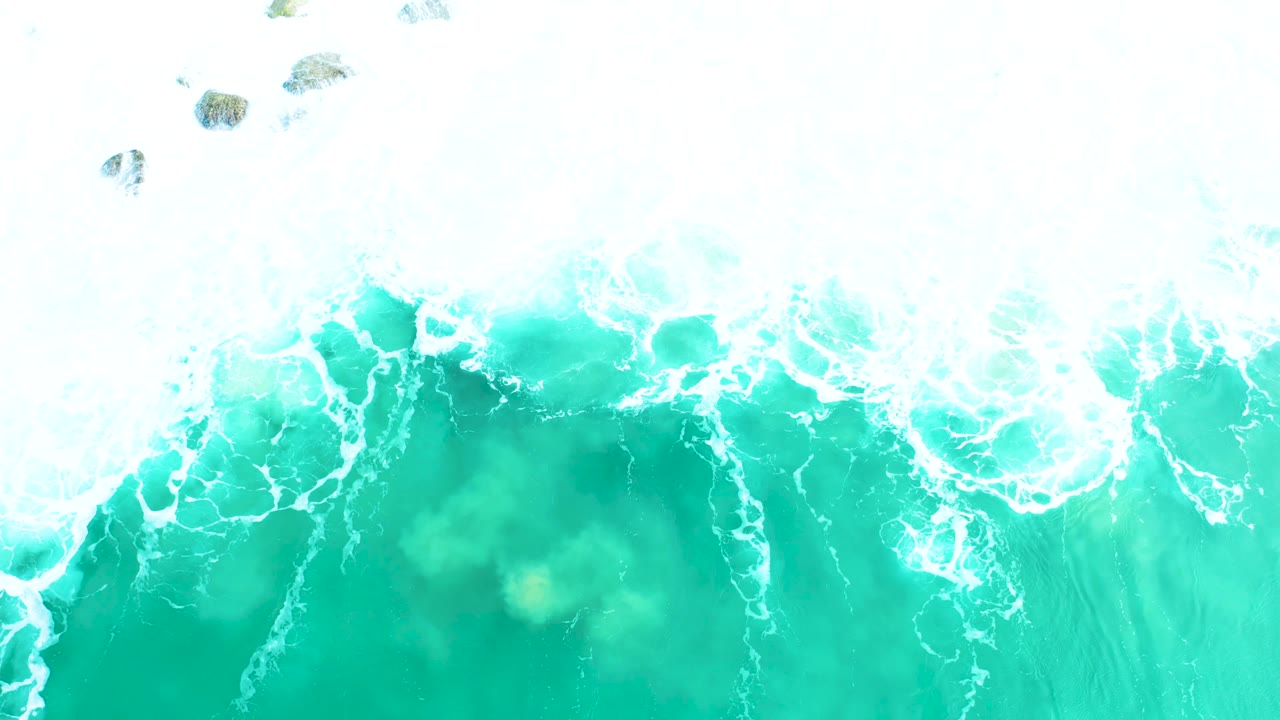 Turquoise surf seen from above with white foam bands and rocks along the upper frame.