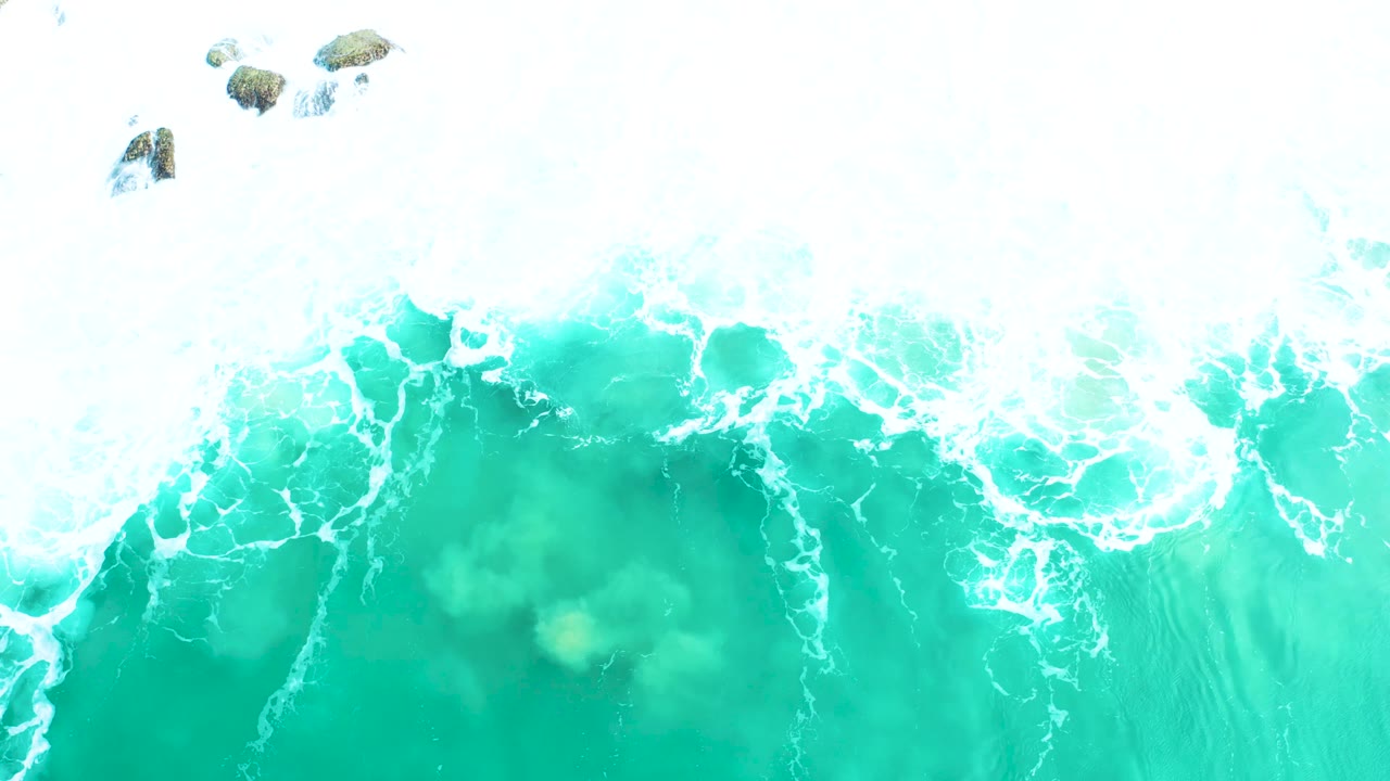 At Marquis Los Cabos, the aerial frame shows turquoise water under a broad white foam edge, with scattered rocks near the top and no visible people.