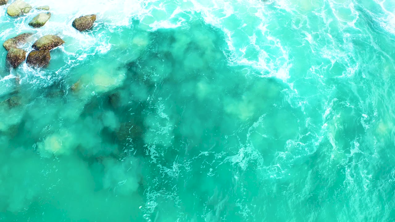 Top-down view of turquoise water and white foam around a small rock cluster at Marquis Los Cabos, with no visible people.