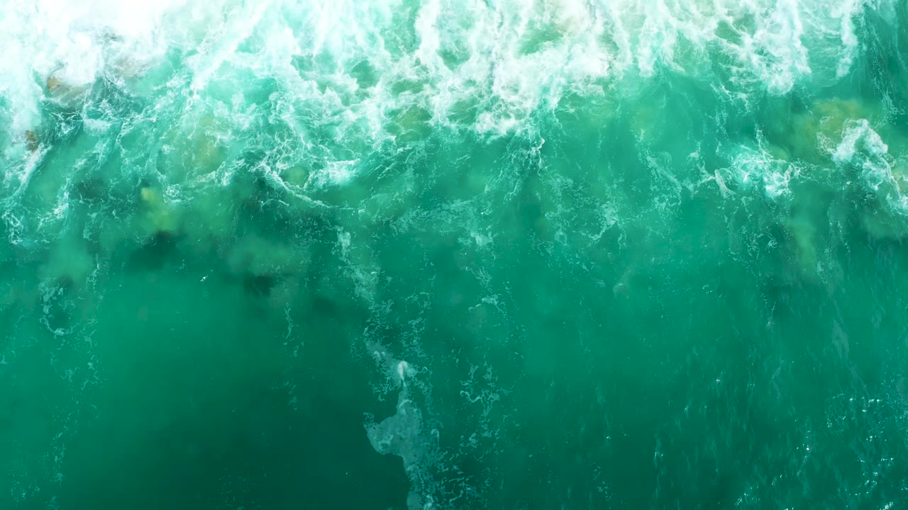 Overhead Marquis Los Cabos frame with foamy wave wash crossing the upper water and dark underwater patches visible through the turquoise surface, with no visible people.
