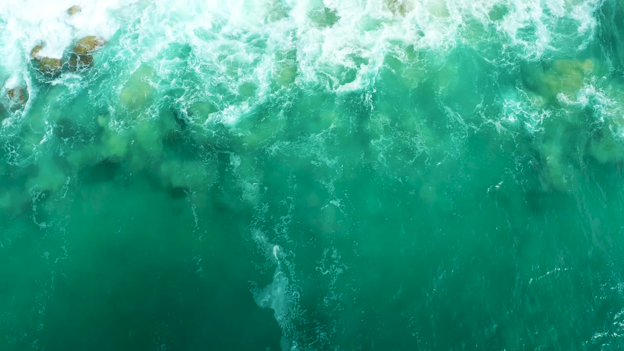 Aerial water view at Marquis Los Cabos with white foam curling across the upper edge, small tan rock shapes near the top, and no visible people.