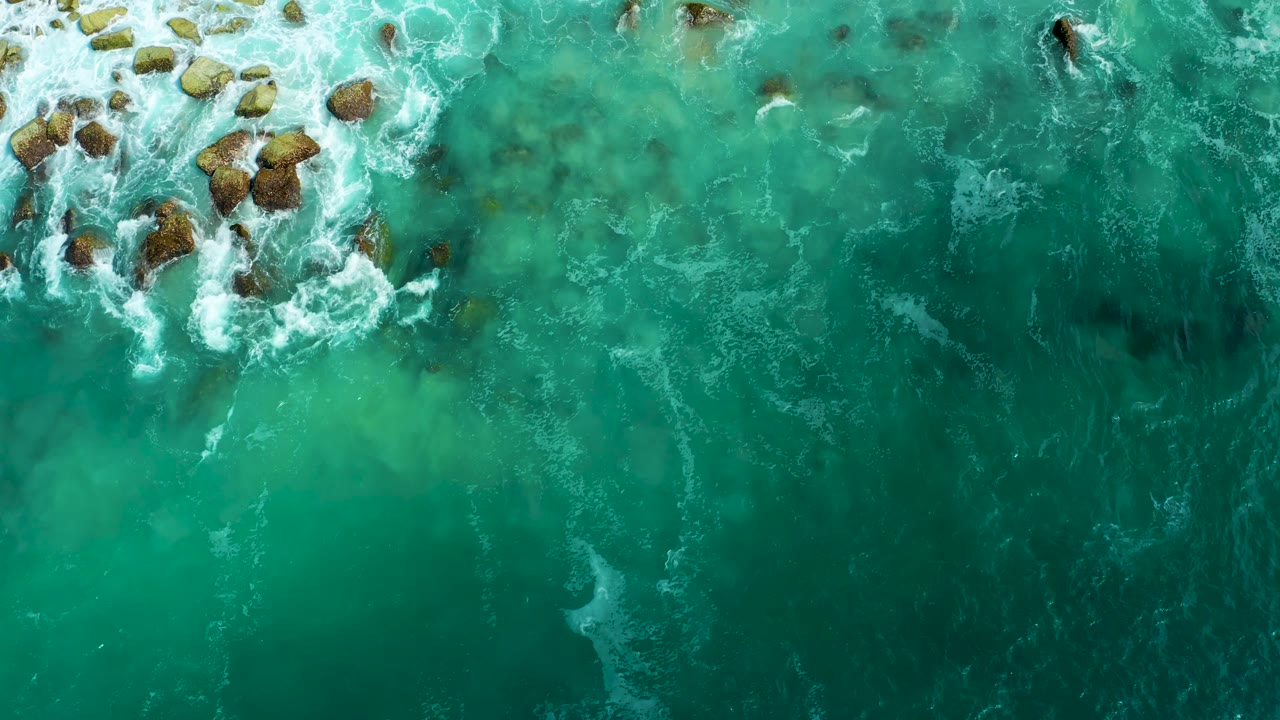 Clear green-blue water fills the overhead frame at Marquis Los Cabos, with clustered rocks near the upper edge and thin white surf lines, no visible people.