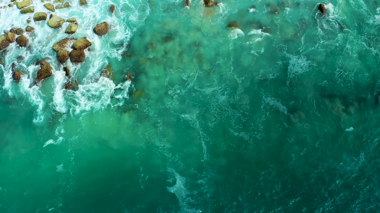 Turquoise water and submerged darker patches surround a small rock cluster at Marquis Los Cabos, with more white foam beginning to wrap the stones and no visible people.