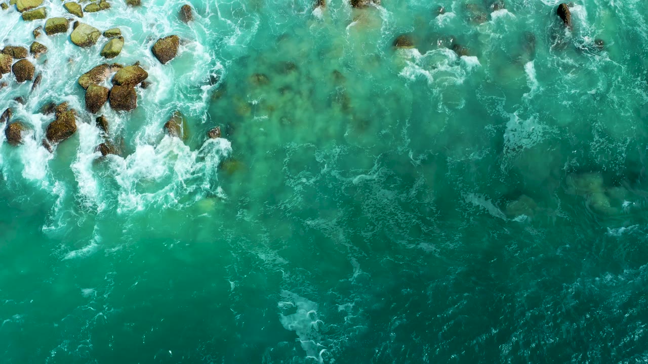 White surf spreads around exposed rocks in the clear turquoise water at Marquis Los Cabos, with no shoreline buildings, boats, or visible people in frame.