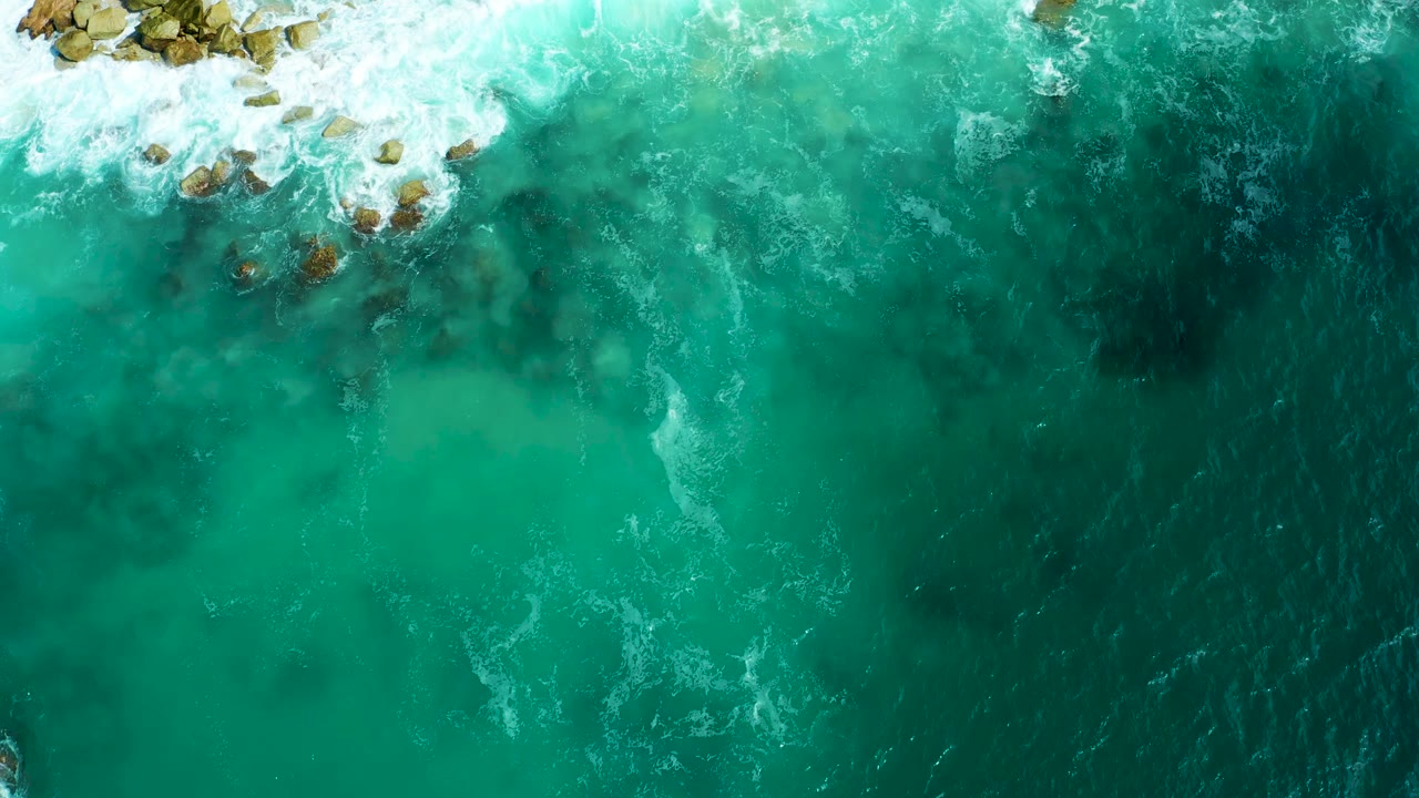 Overhead view of turquoise and deep blue water at Marquis Los Cabos, with white surf breaking around rocks near the top edge and no visible people.