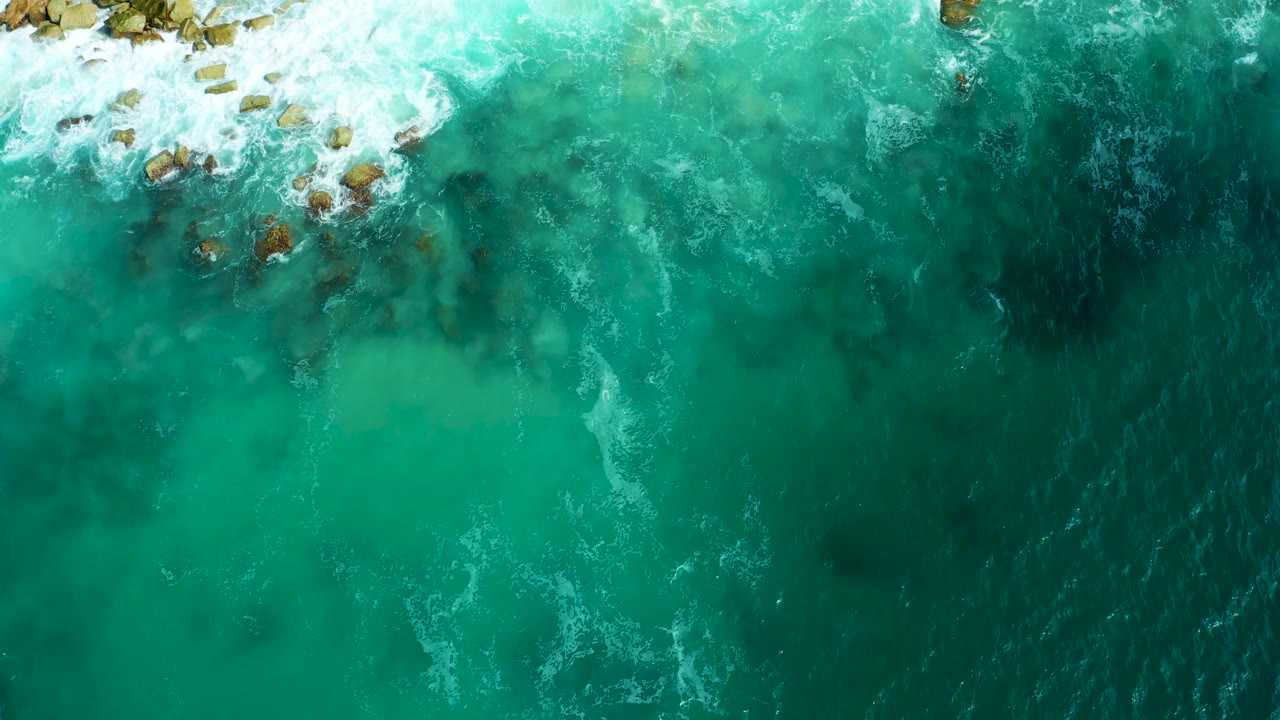Top-down view at Marquis Los Cabos with breaking white water around upper-left rocks and a broad field of green-blue ocean below, with no visible people.