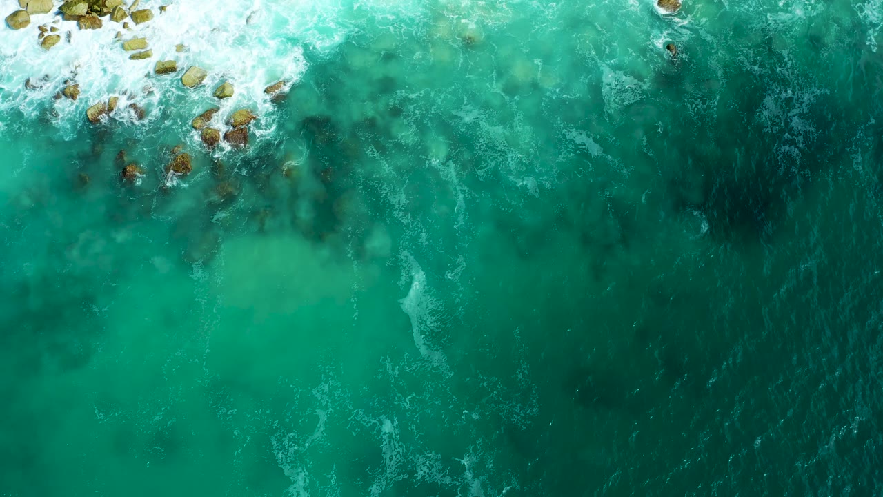Foamy waves break over a rocky shoreline in clear turquoise water.