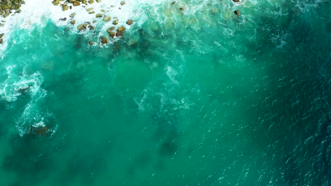 Top-down aerial view of turquoise water and white surf breaking over rocks at Marquis Los Cabos, Mexico, with no visible people.