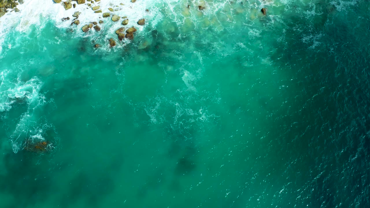 Overhead view of Marquis Los Cabos surf with rocks along the upper edge, milky foam in turquoise water, and a darker open-water band on the right, with no visible people.