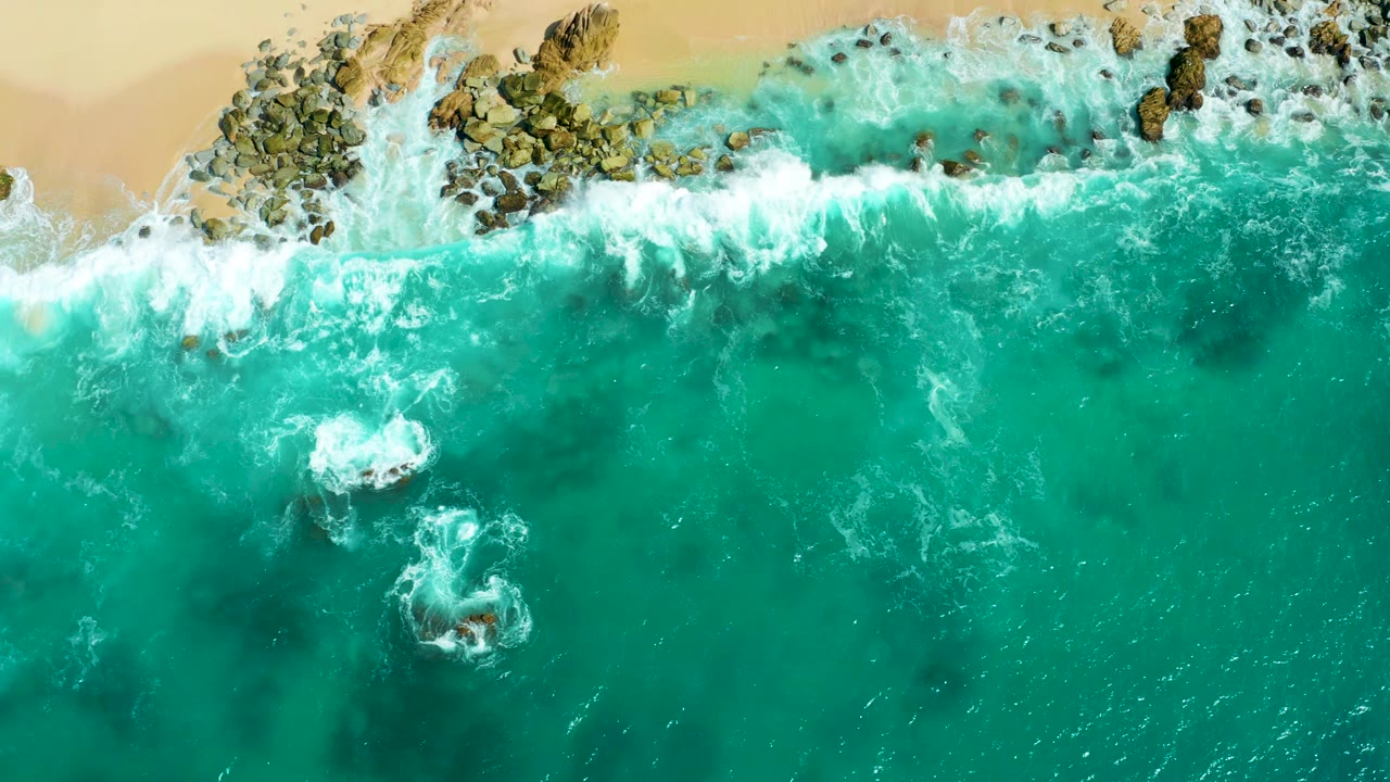 Overhead turquoise surf breaking on rocks beside sandy beach.