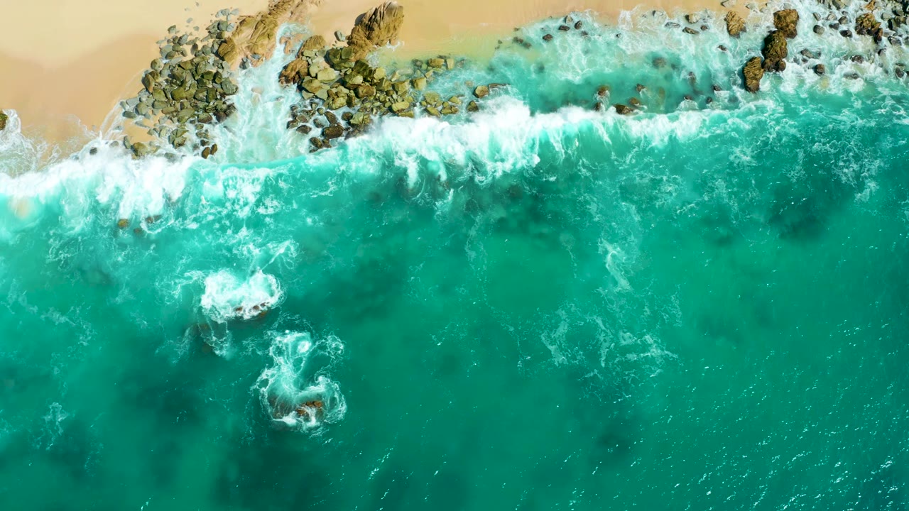 Aerial frame from Marquis Los Cabos with foamy surf spread along the upper shoreline, dark rocks under turquoise water, and no visible people.
