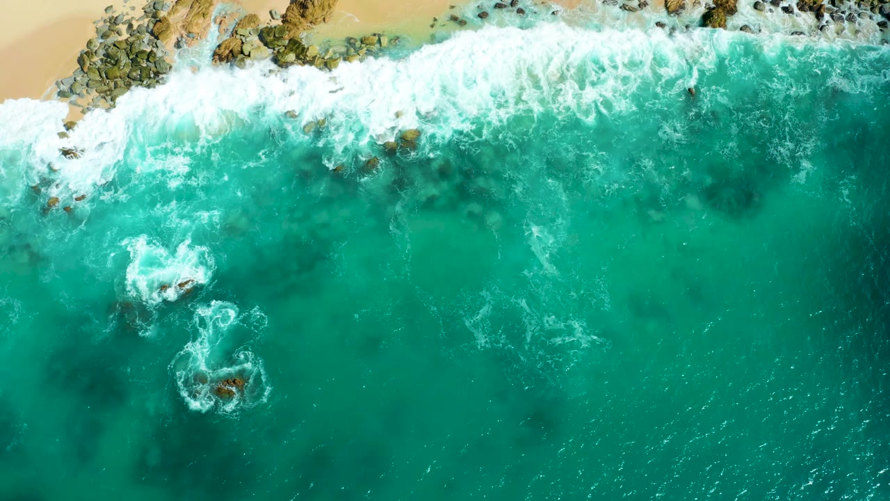 Overhead frame at Marquis Los Cabos with foamy surf spread over the upper rocks, sandy shoreline partly visible, and clear turquoise water below with no visible people.