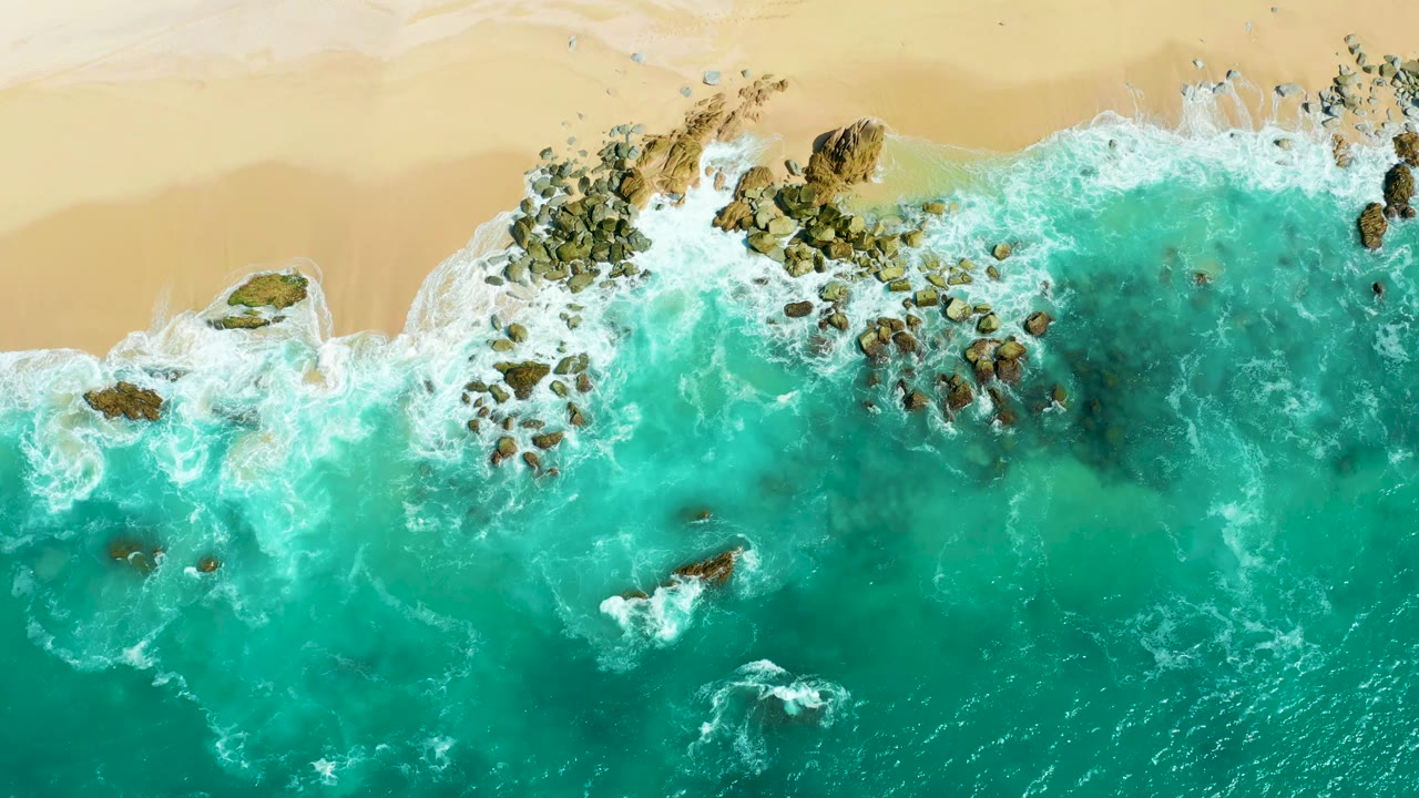 Aerial frame at Marquis Los Cabos where white surf spreads around a central cluster of rocks near the tan beach, with open turquoise water below and no visible people.