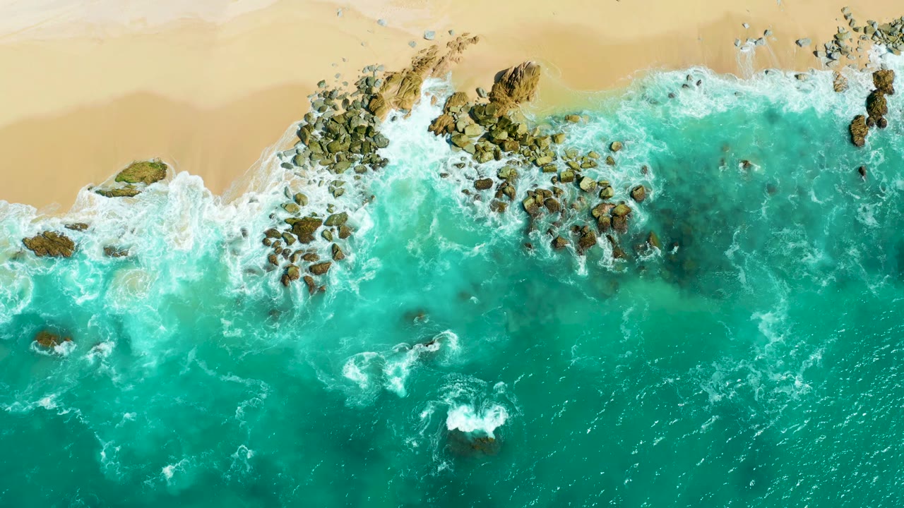 Overhead frame at Marquis Los Cabos showing foam gathering around brown rocks while turquoise water pushes toward the tan sand, with no visible people.