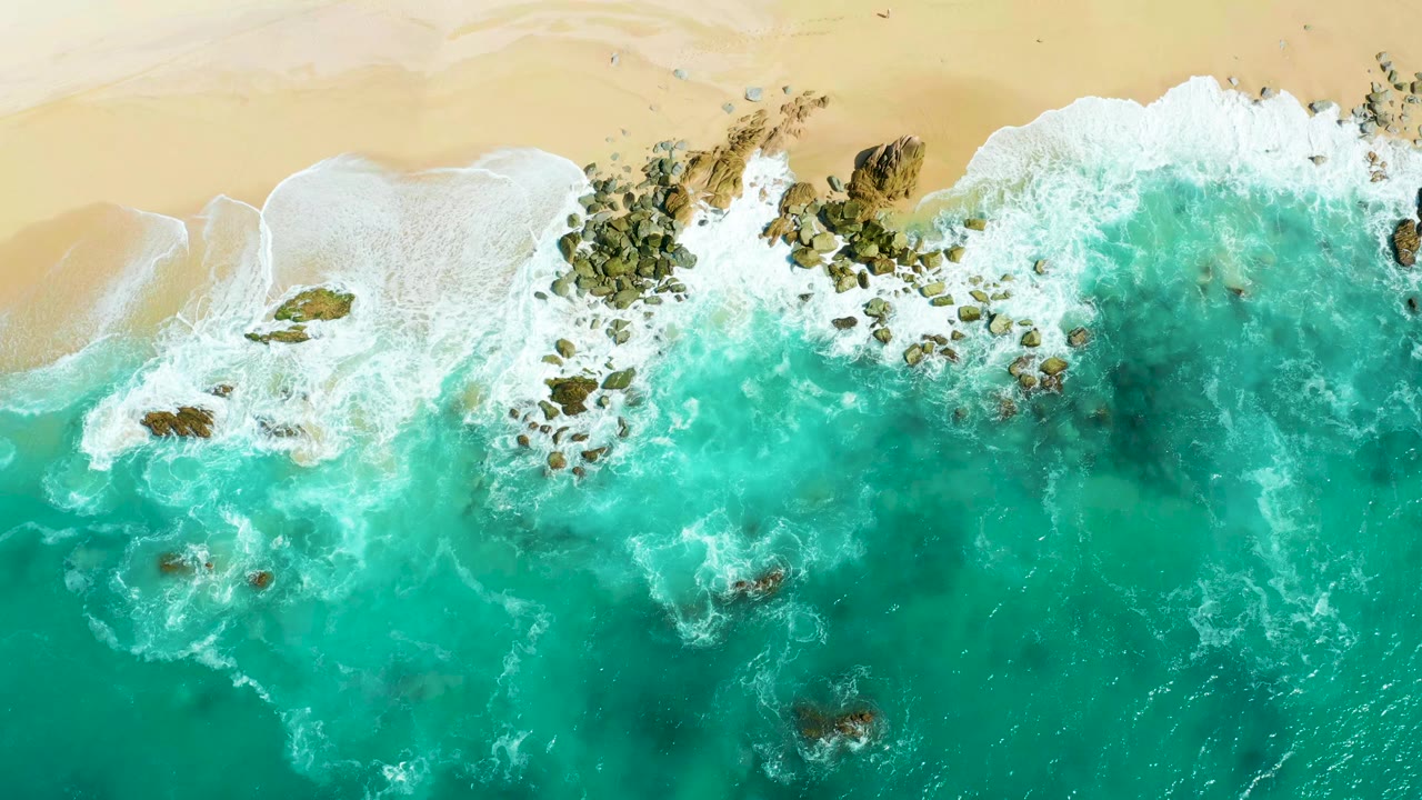High overhead view at Marquis Los Cabos showing turquoise water, white foam, dark rocks, and pale sand along the top edge, with no visible people.