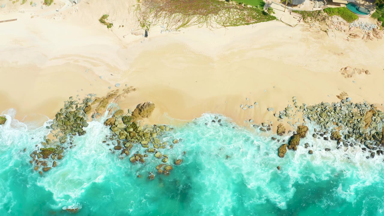 White foam spreads around shoreline rocks below a sandy beach.