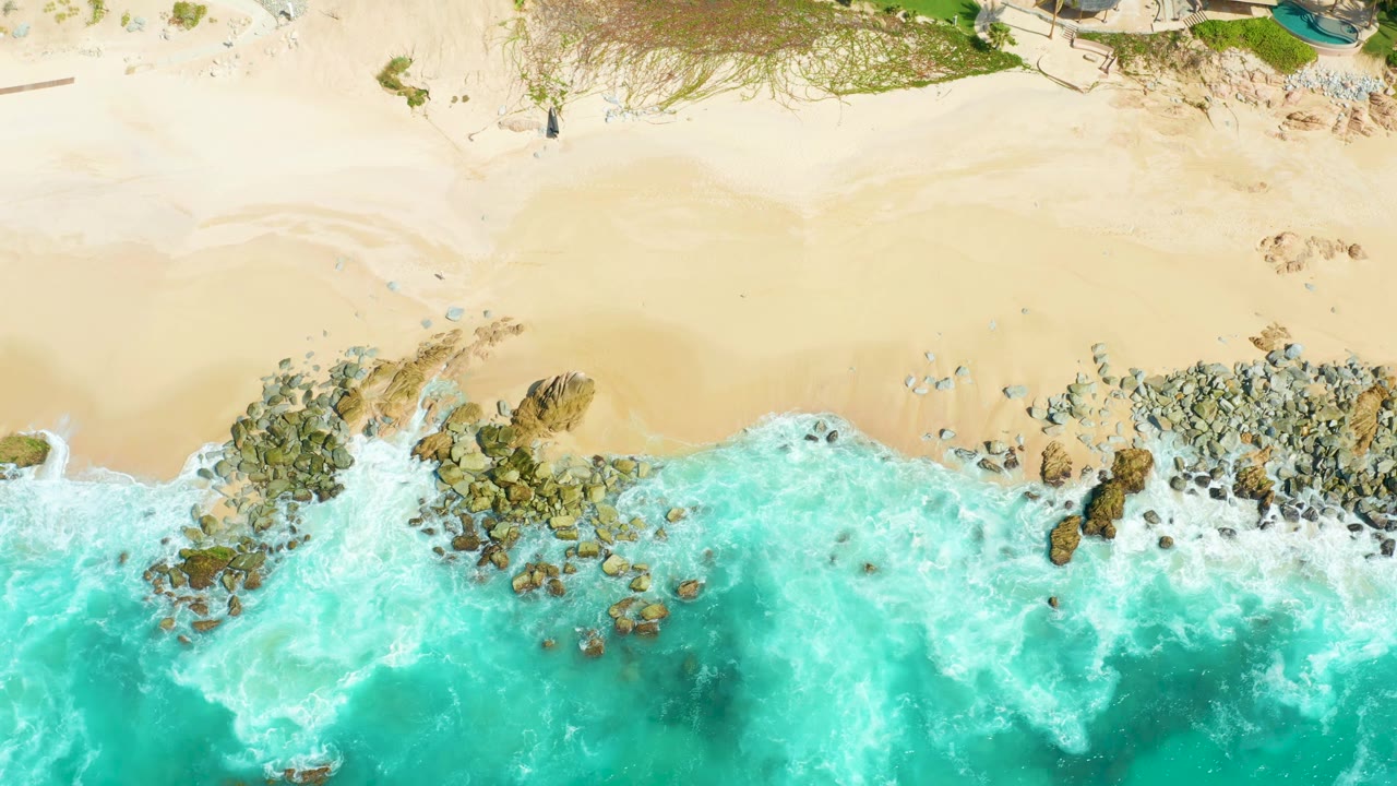 Top-down Marquis Los Cabos shoreline view with turquoise waves pressing into the rocks, pale sand above, and no visible people on the beach.