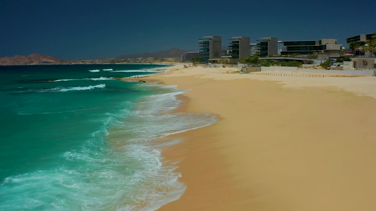 At 0:01, white foam spreads wider along the Dreams Los Cabos shoreline while the turquoise water and beachfront buildings remain in the same elevated view, with no visible people.