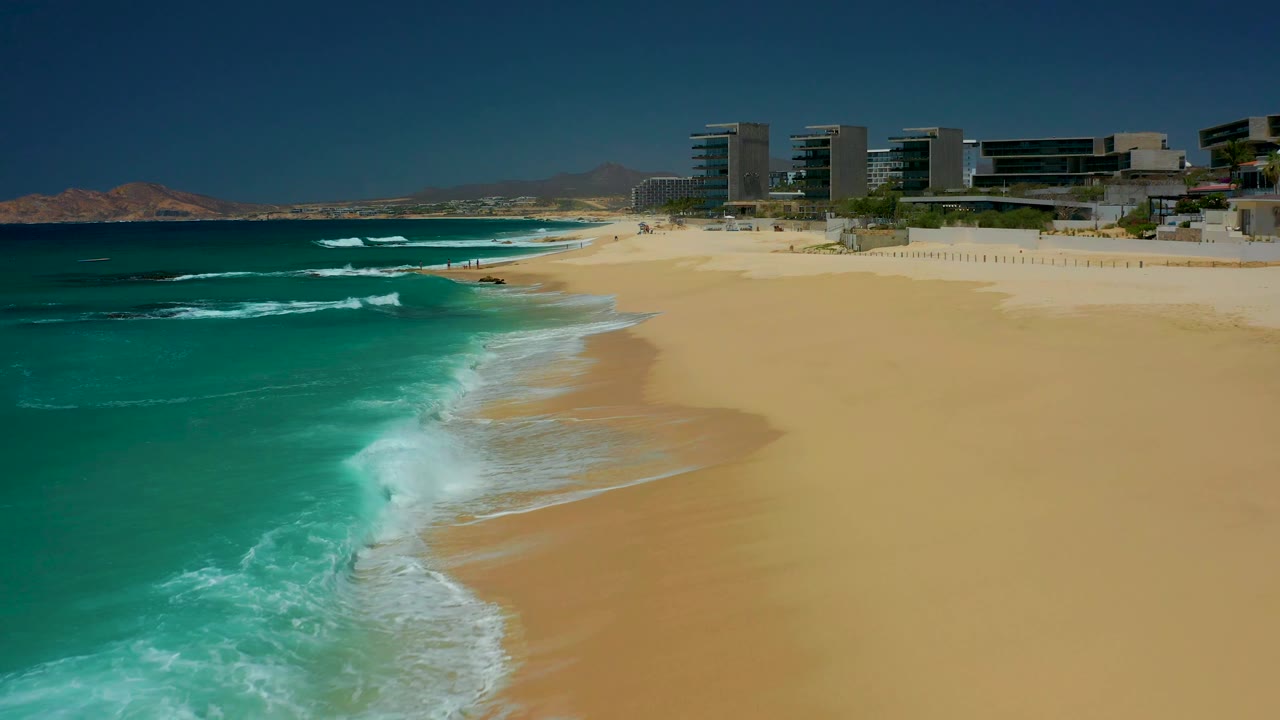 At Dreams Los Cabos, a fresh line of foam reaches into the sandy foreground as the resort buildings sit high on the beach and no people are visible.